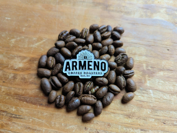 Enamel pin of the Armeno Coffee Roaster's "bean sunrise" Logo displayed on a pile of coffee beans