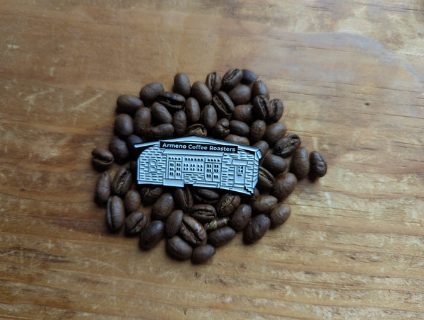 Enamel pin of The Mill at Smith pond, aka Armeno Coffee Roaster's location, displayed on a pile of coffee beans.